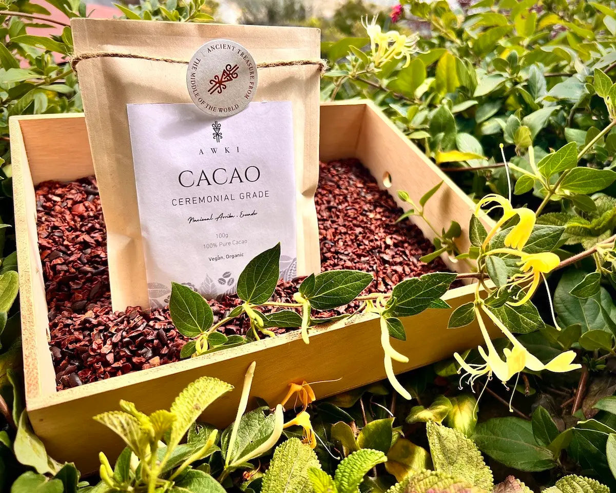 Awki ceremonial cacao 100g pouch placed in a wooden tray filled with cacao nibs and surrounded by fresh green plants