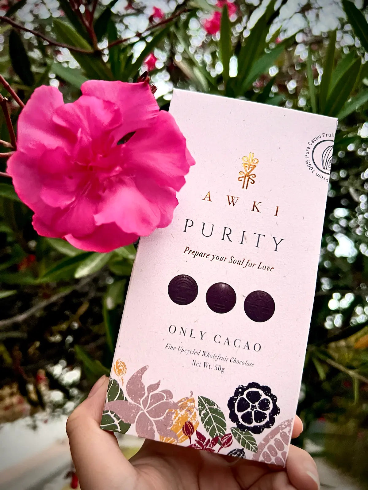 Awki Purity cacao bar 50g held next to a vibrant pink flower outdoors