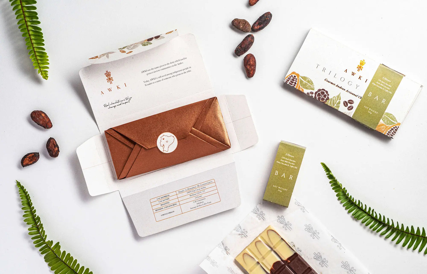 Awki Trilogy Bar unboxing with gold inner wrap, cacao beans, and three-flavor chocolate bar on white background with ferns