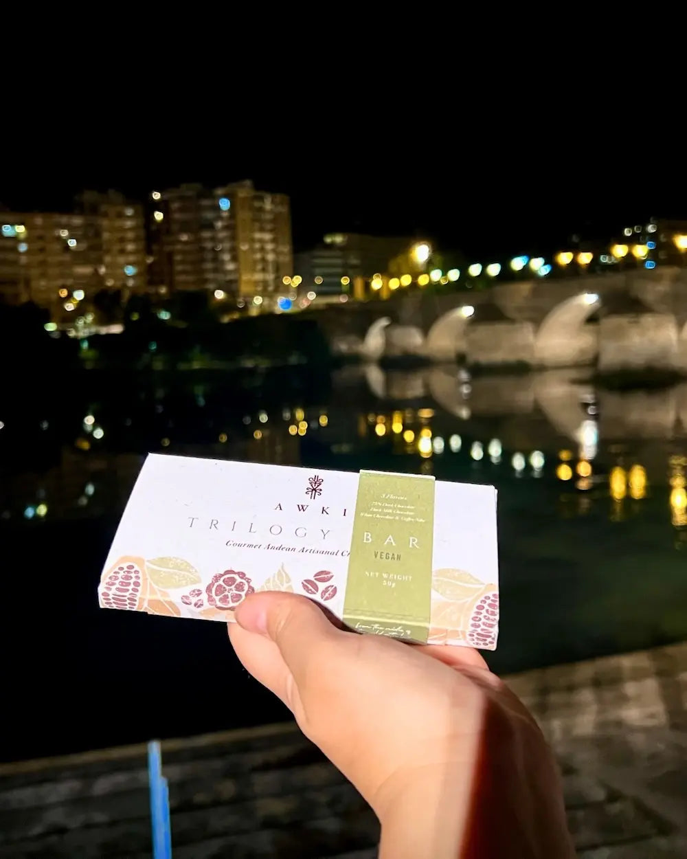 Awki Trilogy chocolate bar held at night with city lights and a historic stone bridge in the background