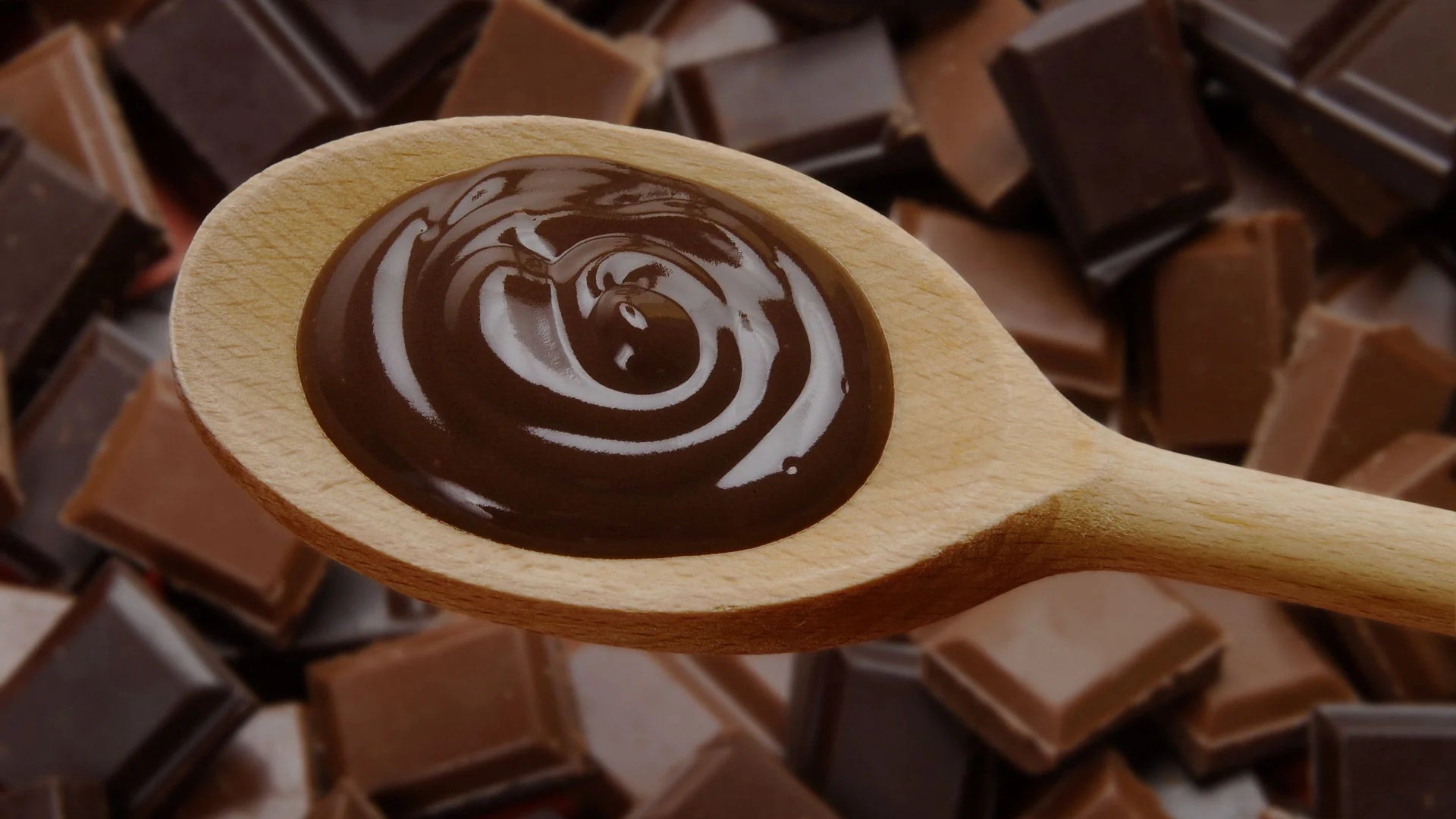 Swirl of melted dark chocolate on a wooden spoon with chocolate chunks in the background